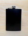 A matte black metal flask with a silver screw cap and attached clip, standing upright on a light wood surface against a matching wood background.