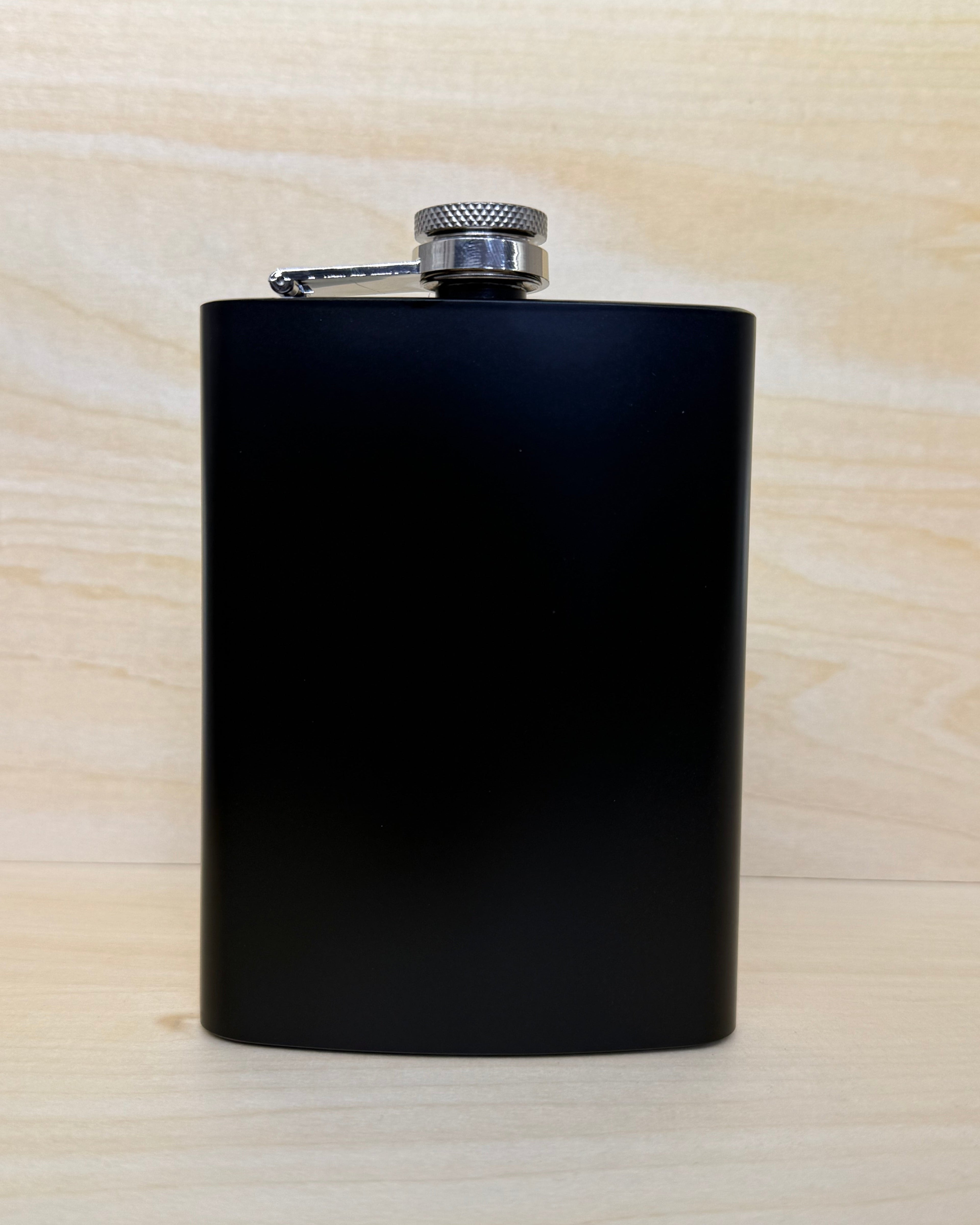 A matte black metal flask with a silver screw cap and attached clip, standing upright on a light wood surface against a matching wood background.