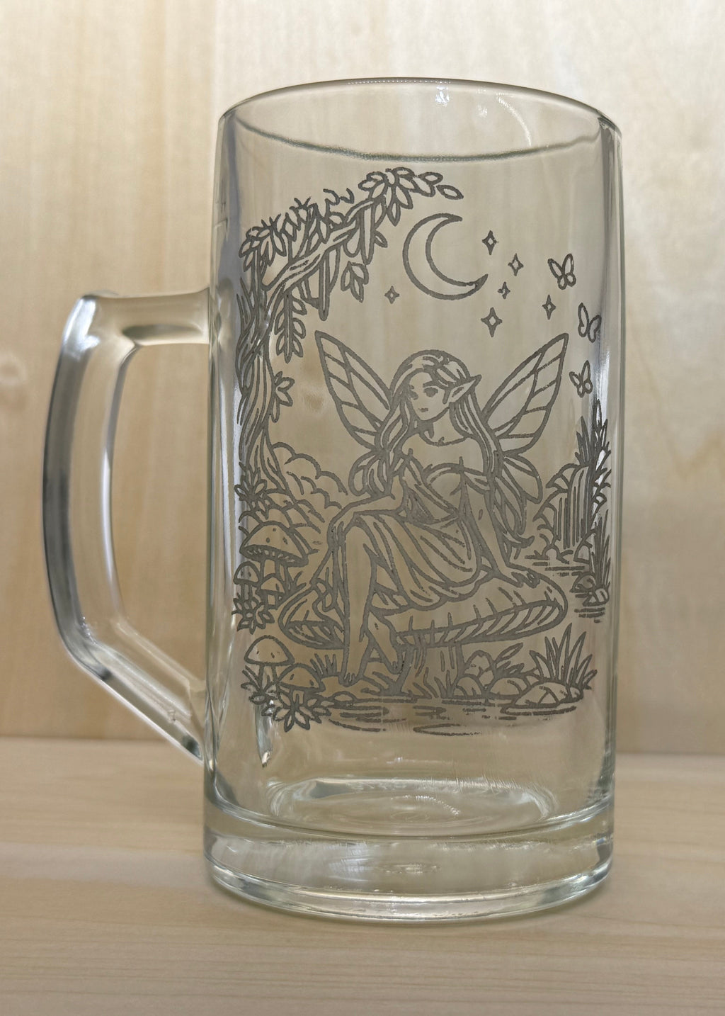 Glass mug with engraved fairy design on a wooden surface