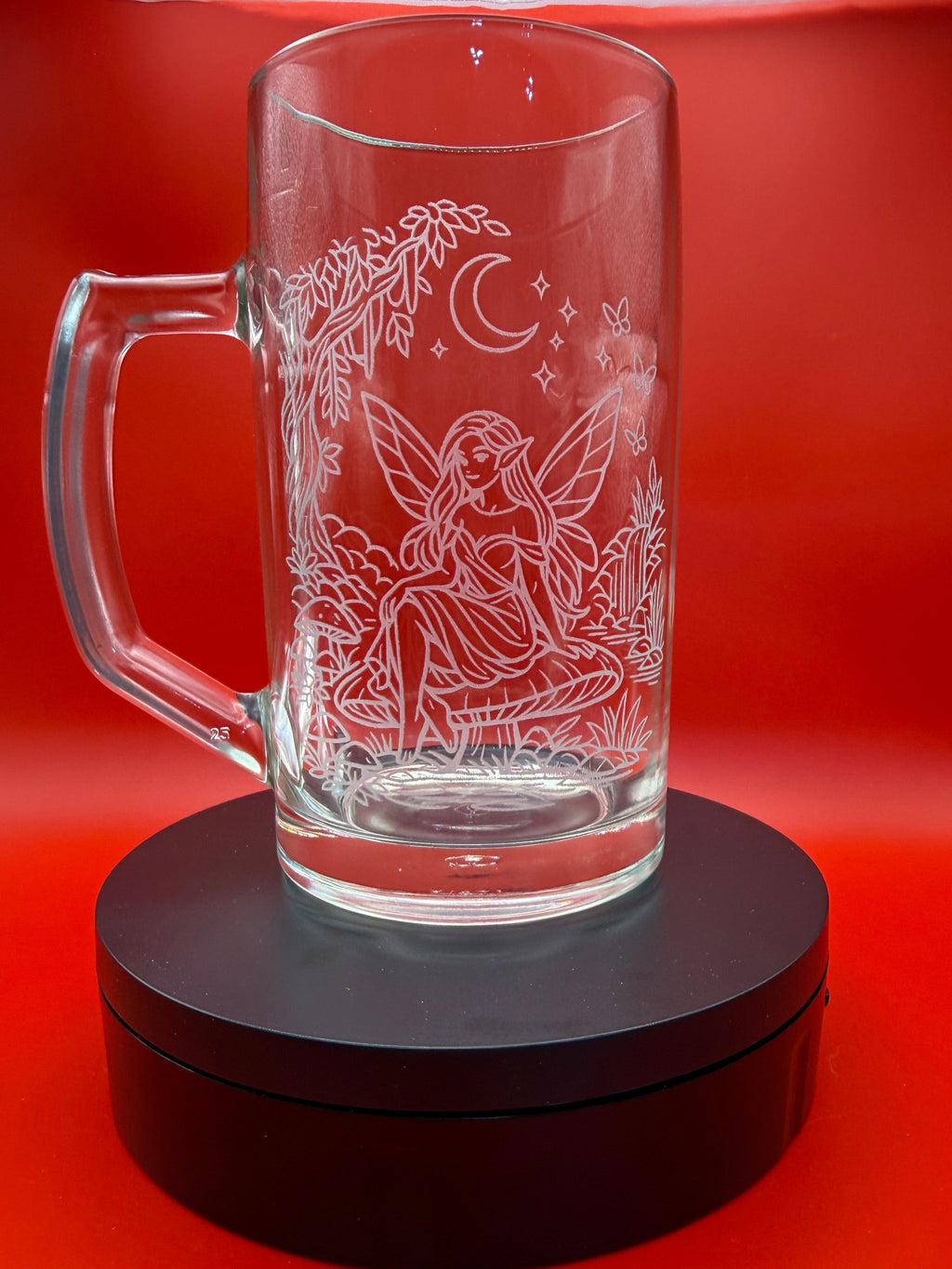 A clear glass mug with a handle, featuring an etched fairy, crescent moon, and stars design, displayed on a black circular stand against a vibrant red background.