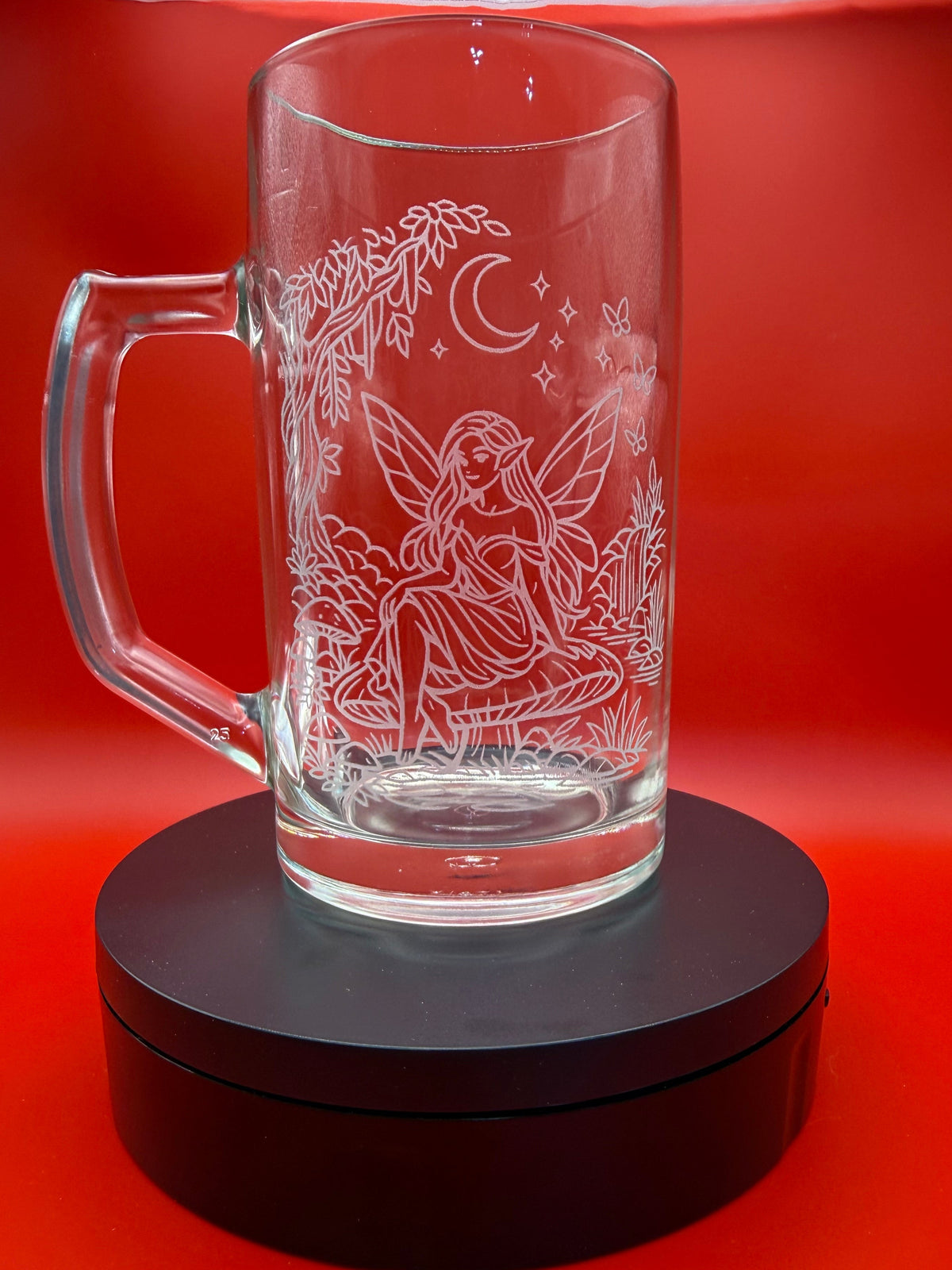 A clear glass mug with a handle, featuring an etched fairy, crescent moon, and stars design, displayed on a black circular stand against a vibrant red background.