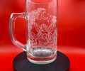 A clear glass beer mug with a handle, featuring white etched line art of a fairy under a crescent moon, displayed on a black circular base against a vibrant red background.