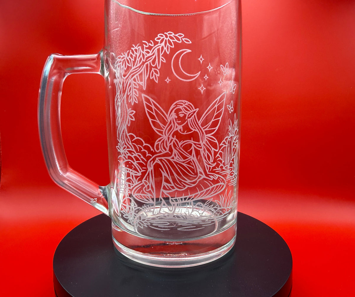 A clear glass beer mug with a handle, featuring white etched line art of a fairy under a crescent moon, displayed on a black circular base against a vibrant red background.
